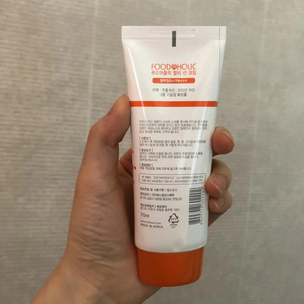 A hand holding a white tube of FOODFOHOLIC hand wash with orange accents, showing the back of the packaging with Korean text and a barcode, against a neutral gray textured background.