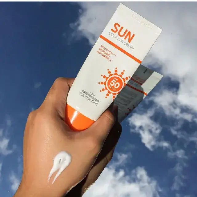 A hand holding a white and orange tube of SUN Safe Sun Cream SPF 50 against a blue sky with clouds, with white sunscreen cream visible on the back of the hand.